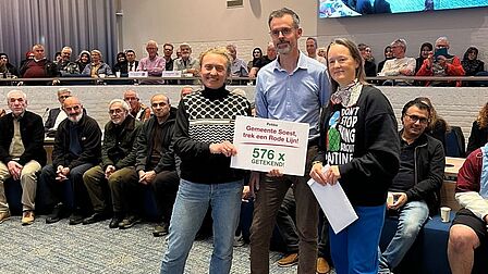Foto van drie mensen die samen een document vasthouden, met op de achtergrond tientallen inwoners.