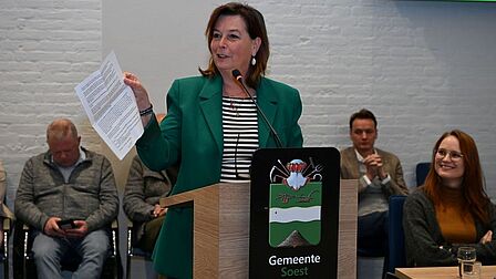 Foto van Tineke Flinterman die een vel papier omhoog houdt en lachend spreekt.
