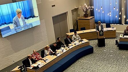 Foto van de raadzaal met zeven raadsleden en een man achter een spreekgestoelte, op de achtergrond een scherm met daarop de man achter het spreekgestoelte.