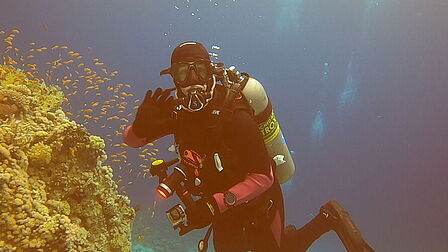 Foto van Anne onder water in een duikpak met duikfles op haar rug bij een groot koraal met ene school kleine vissen.