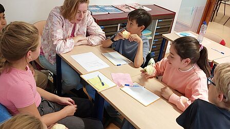 Foto van kinderen en een vrouw rond een schooltafel.